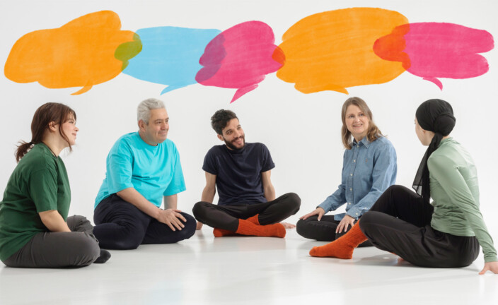 Five people sit on the floor talking. Speech bubbles of different colors have been added to the image in the form of cartoons.