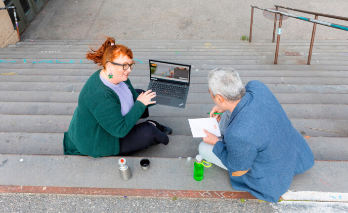 Nainen ja mies istuvat portailla ja keskustelevat. Naisella on sylissään kannettava tietokone, jonka näytöllä näkyvää sisältöä hän esittelee miehelle. Miehellä on käsissään kynä ja muistilehtiö.